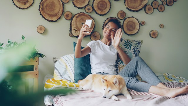 Woman using a smartphone for a video call while a dog lies beside her on a bed.