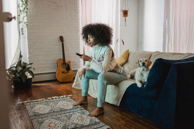 Woman sitting on a sofa with a dog beside her while holding a television remote.